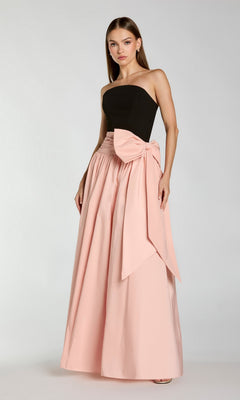 A woman wearing the Long Formal Dress: Mac Duggal 2370 in Black Blush, featuring a strapless black top and a long, high-waisted pale pink skirt with an oversized bow on the side, stands against a plain light background.
