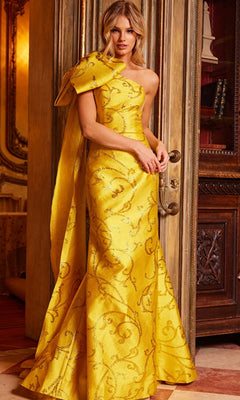 Woman wearing the Jovani One-Shoulder-Bow Long Formal Dress 23742 in yellow with an ornate pattern and large bow detail, standing indoors by a decorative door.
