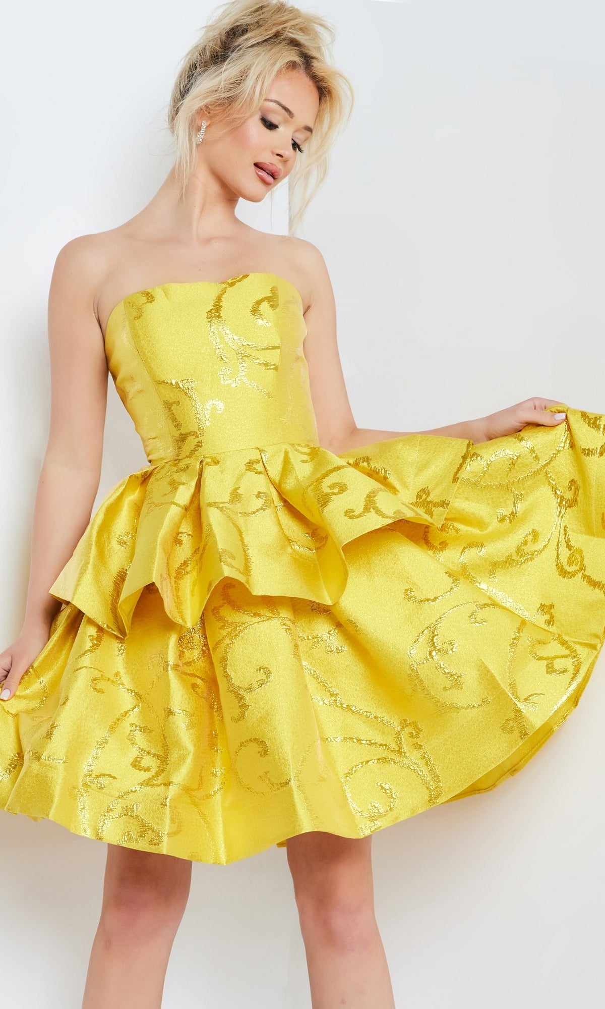 A woman models the Jovani Short Yellow Strapless Party Dress 23743, featuring ruffles and an ornate shiny pattern, as she holds out the skirt against a plain light background.