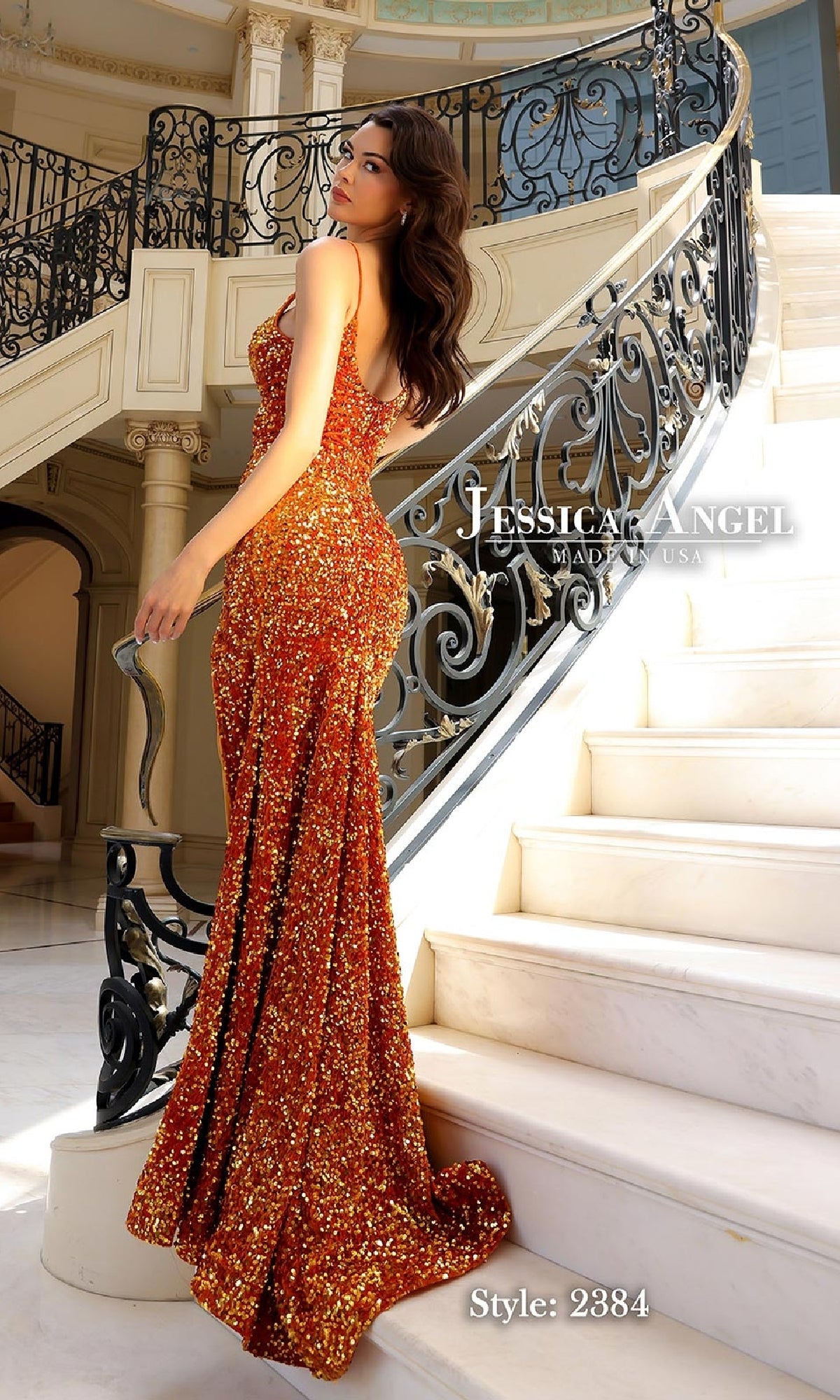 A woman in a shimmering orange Jessica Angel 2384 long sequin velvet prom dress with a train stands on marble stairs in an elegant interior.
