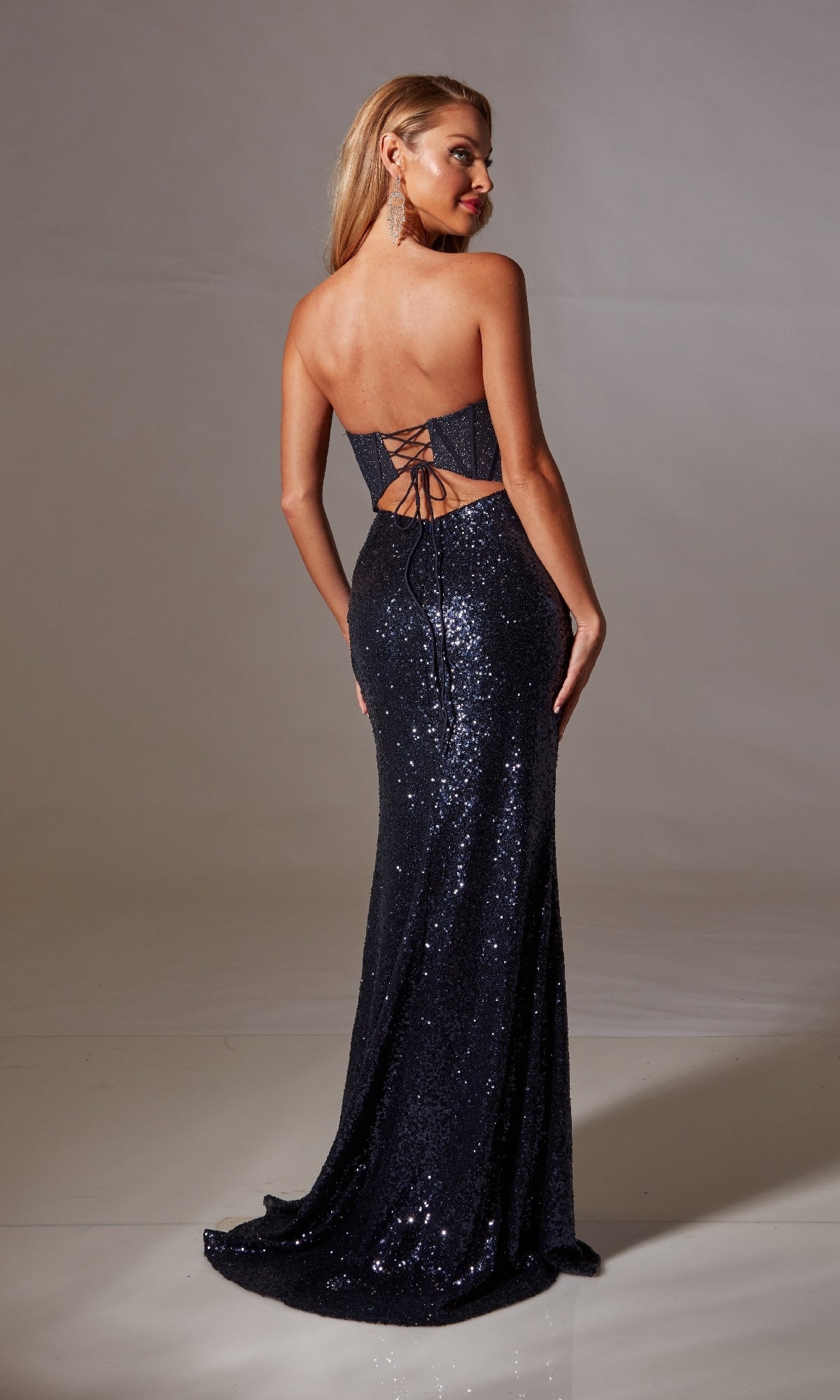 A woman wearing the Aerie Couture 238 Strapless Sequin Long Prom Dress, featuring a corset bodice and lace-up open back, stands looking over her shoulder.