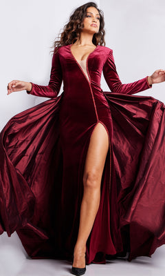 A woman stands gracefully against a plain background wearing the Jovani Long Sleeve Long Velvet Formal Dress 23943, a deep red evening gown featuring a high slit and flared skirt.