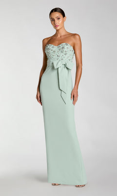 A woman wearing the Mac Duggal 2395 long formal dress in sage, featuring a strapless design with floral embellishments on the bodice and a statement waist bow, stands against a plain background.