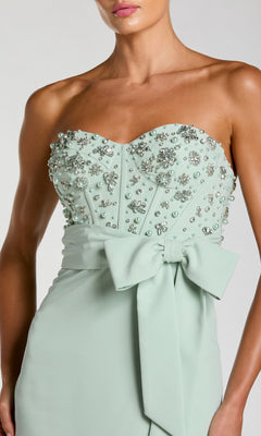 Model wears the Mac Duggal 2395 long formal dress, featuring a strapless, light green design adorned with rhinestone embellishments and a large bow at the waist; shown from shoulders to hips.