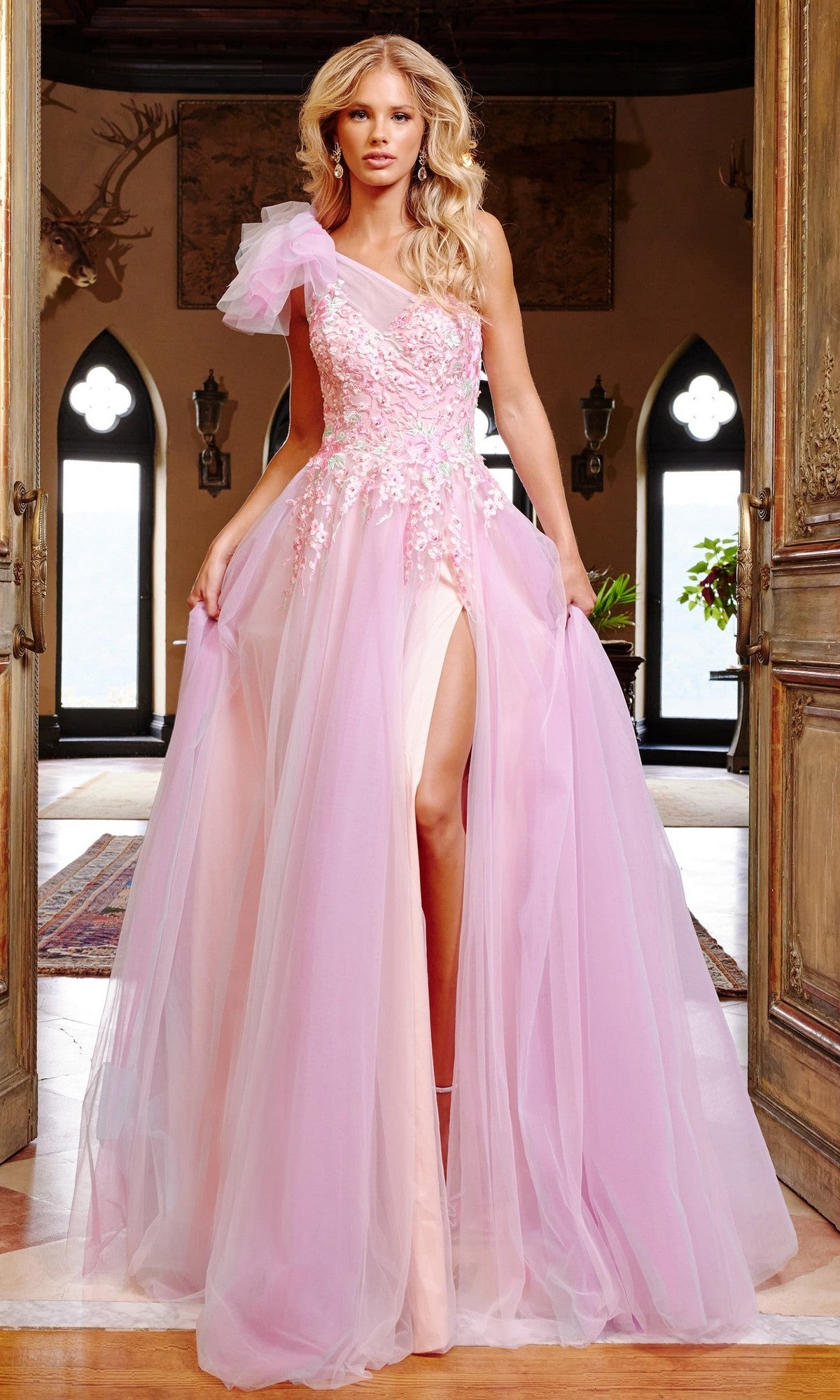 A woman stands indoors wearing the Jovani 23951, a pink multi long prom dress featuring a one-shoulder design, floral embroidery, sheer tulle layers, and a high leg slit.