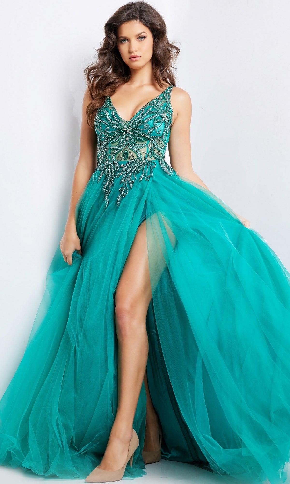 Woman in a Jovani Beaded-Bodice Long Prom Ball Gown 23962, featuring a teal beaded bodice, deep V-neckline, tulle skirt with high slit, and nude heels, standing against a plain background.