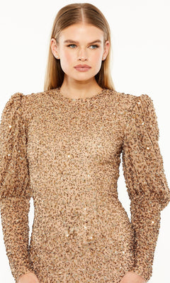 A woman models the Long Formal Dress 24000 by Mac Duggal, featuring shimmering gold sequins, puffed shoulders, and long sleeves, set against a white background.