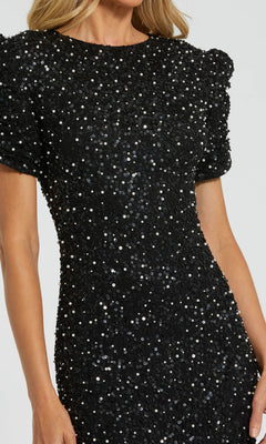 A person wears the Mac Duggal 24003 short sleeve sequin formal dress, its fitted black silhouette sparkling against a plain light background.