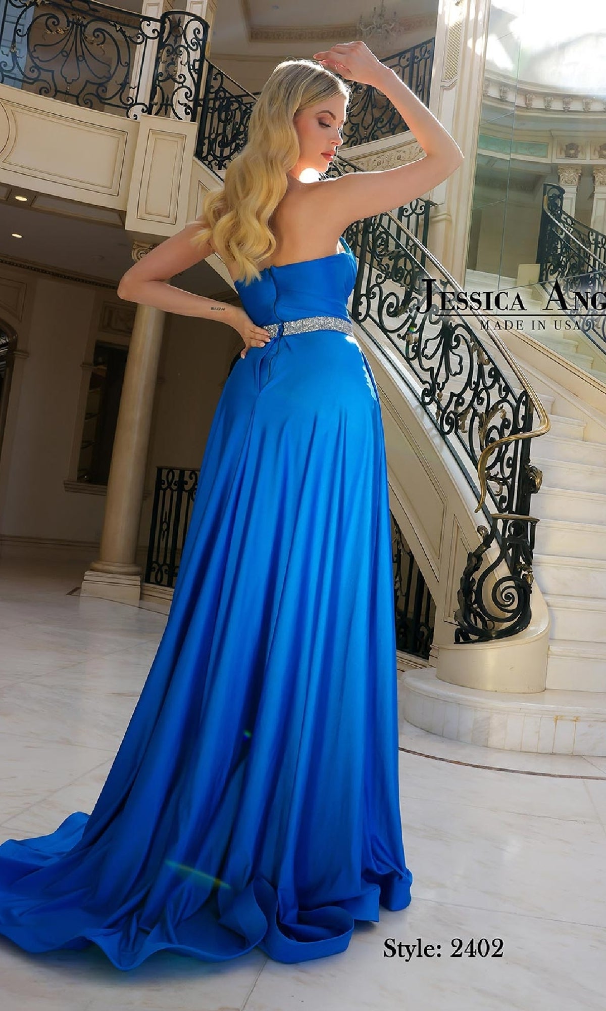 A woman in a strapless high-low prom dress, Jessica Angel 2402, with a jeweled waistband stands in a marble foyer near a staircase. The royal blue design exudes elegance. 