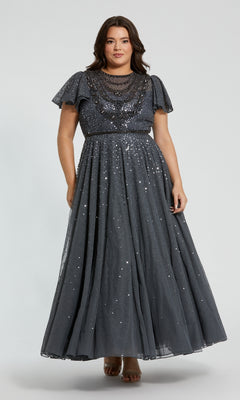A woman wears the Plus-Size Charcoal Formal Dress: Mac Duggal 24025, a floor-length, short-sleeve evening gown embellished with sequins and beading, standing against a plain light background.