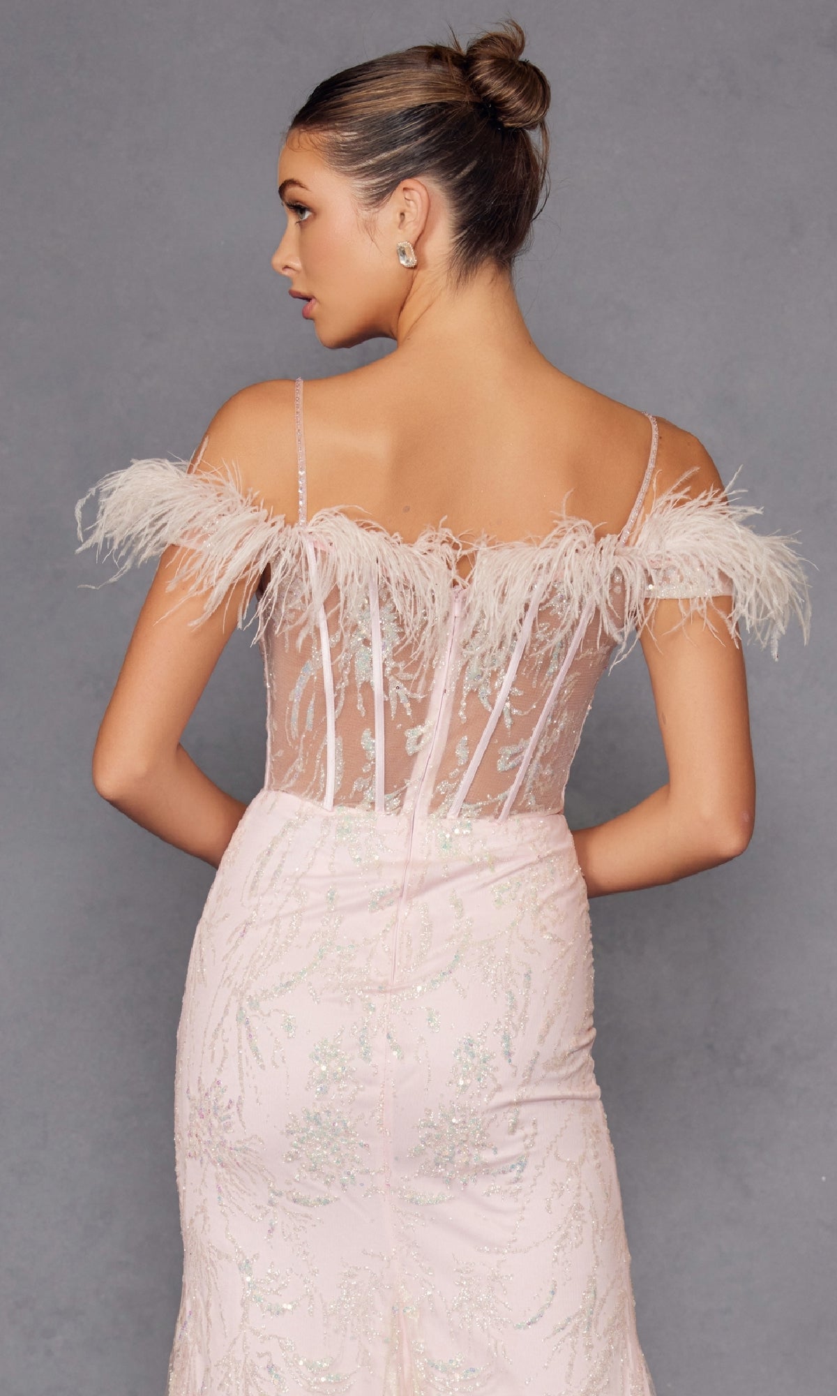 A woman with a bun is seen from the back wearing the Feather Cold-Shoulder Long Glitter Prom Dress 2402, a light pink gown with sequin embellishments and feather details, shown against a gray background.