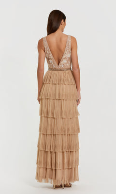 A woman stands facing away, wearing the Mac Duggal 24061 Ruffled Long Nude Formal Dress—a sleeveless gown with a fitted bodice, deep V-back, and a tiered fringed skirt in light beige.