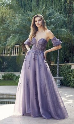 A woman poses outdoors in the Puff-Sleeve Strapless Glitter Prom Ball Gown 2409, featuring floral embroidery and a sheer, flowing skirt. Lush greenery and a pool in the background highlight her elegant style.