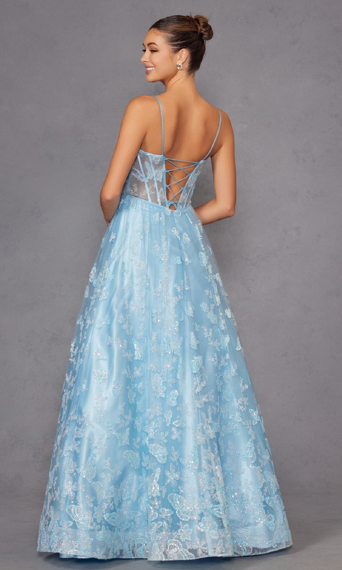 A woman stands against a plain gray background wearing the Sheer-Corset Glitter-Print Long Prom Dress 2413, a light blue, floor-length A-line gown with delicate lace, thin straps, and a lace-up corset back.
