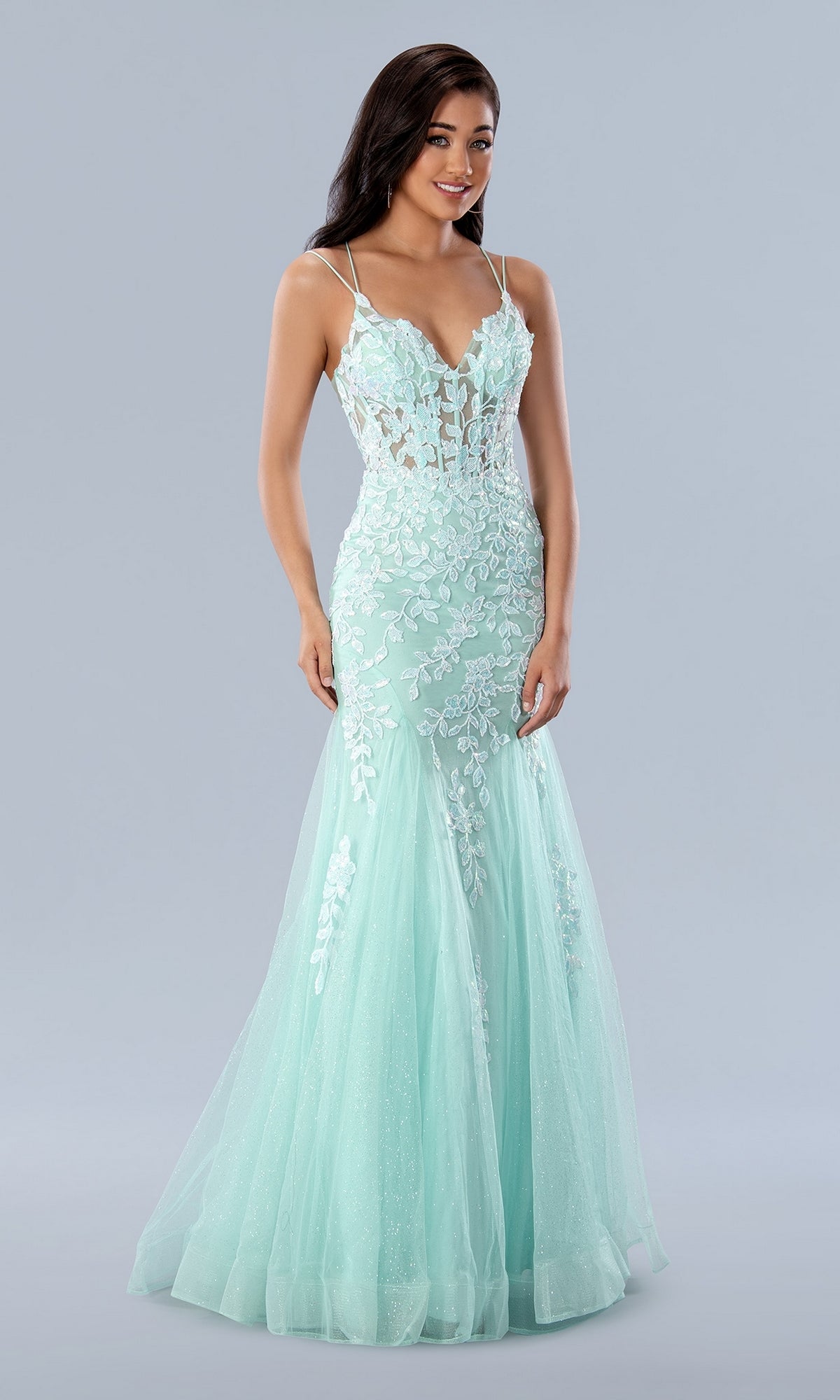 A woman stands against a grey background wearing the Long Prom Dress 24229 by Stella Couture—a light blue, fitted mermaid gown with lace embroidery, a sheer bodice, and thin straps.