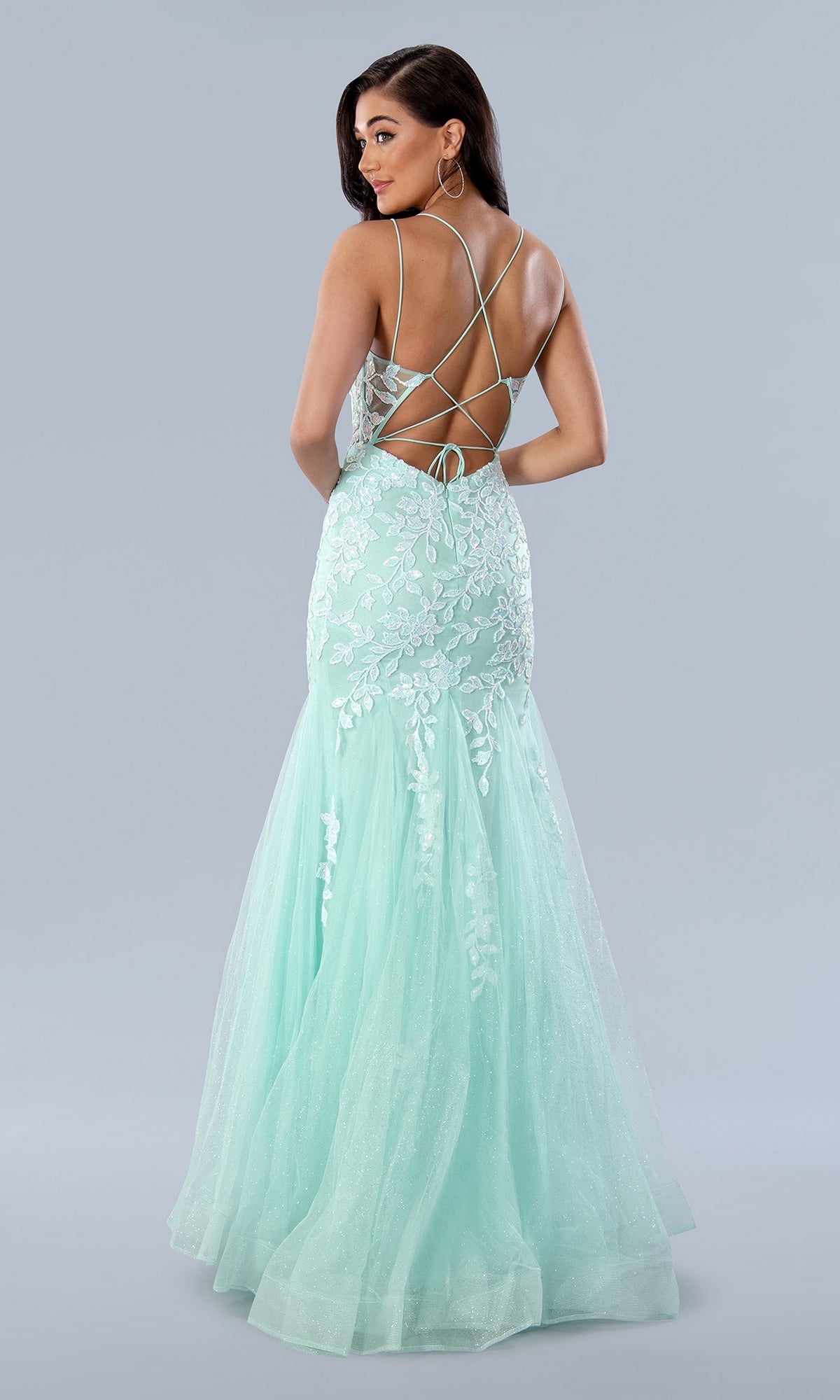 A woman poses in the Long Prom Dress 24229 by Stella Couture, a light blue mermaid gown with lace embroidery and a crisscross open back, set against a plain background.