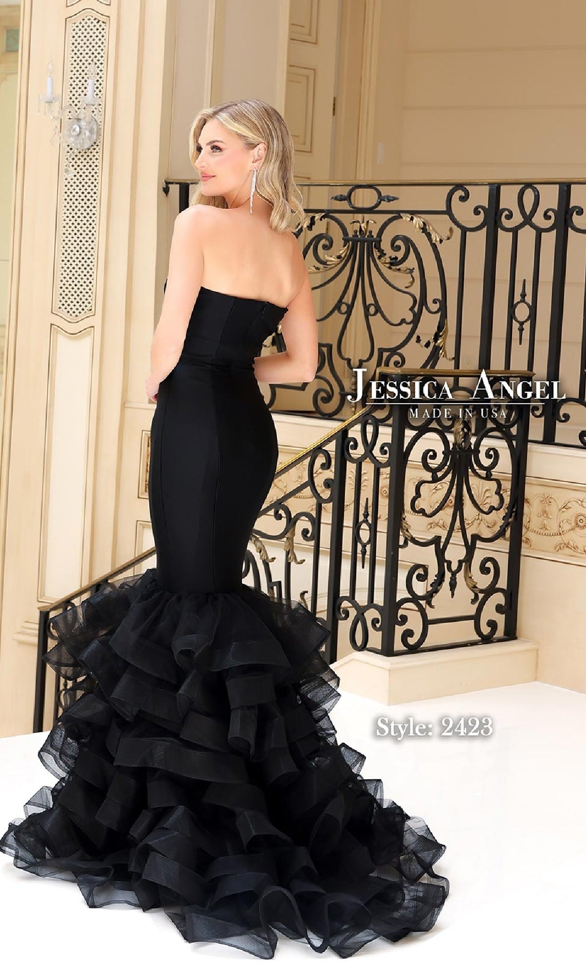 A woman stands by a decorative staircase in the elegant black Strapless Long Prom Dress: Jessica Angel 2423, featuring a mermaid silhouette and layered ruffles.