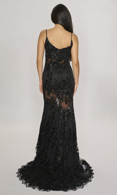 A woman stands facing away, wearing a sleeveless fitted lace-embroidered long dress with sheer floral details, thin straps, and a slight train—the Long Formal Dress: Nina Canacci 2429 in black.