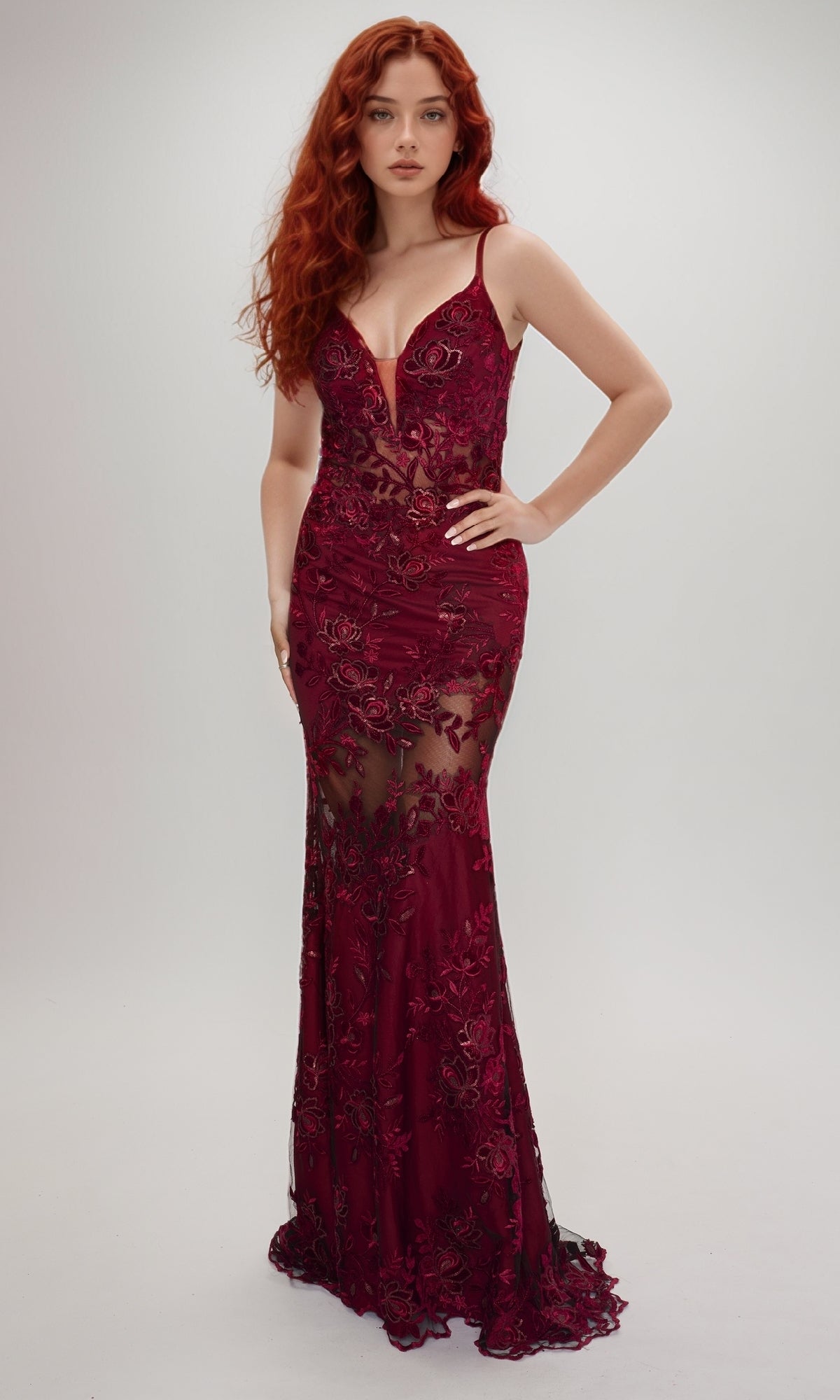 A woman with long red hair wears the Nina Canacci 2429, a sleeveless fitted burgundy gown featuring a deep V-neck, floral lace details, sheer panels, and a floor-length silhouette, standing against a plain light background.