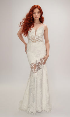 A woman with long red hair wears the Nina Canacci 2429, a floor-length white lace-embroidered formal dress with sheer panels and floral patterns, standing against a plain light background.