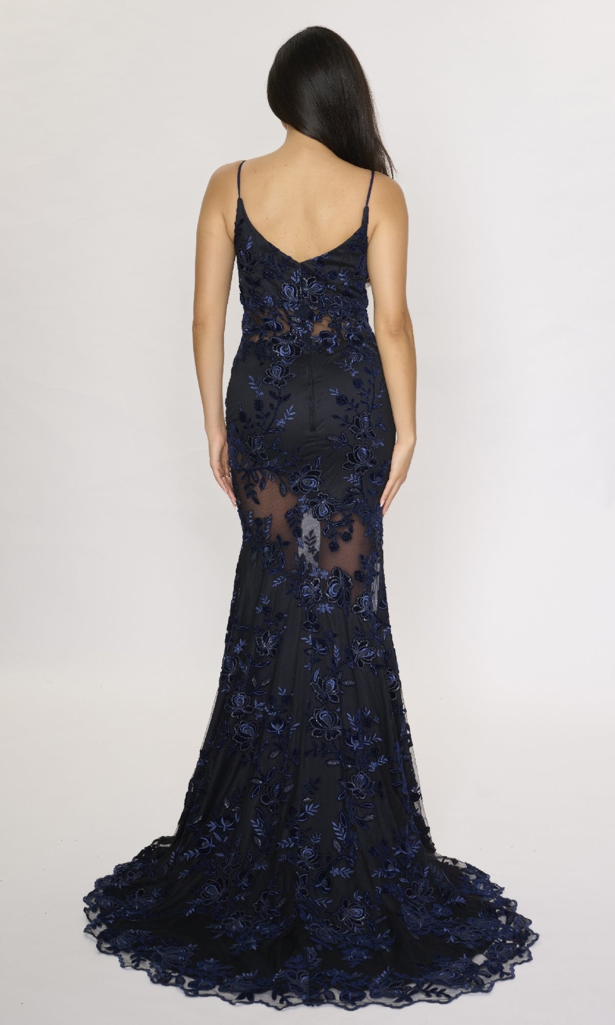 A woman stands with her back to the camera in the Nina Canacci 2429—a navy blue, sleeveless, floor-length formal dress featuring floral lace and a slight train.