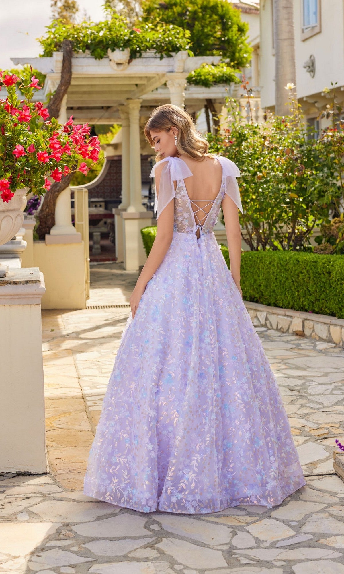 A woman wearing the Juliet JT2439K Lilac Purple Long Lace Prom Dress stands outdoors on a stone path, surrounded by greenery and flowers.