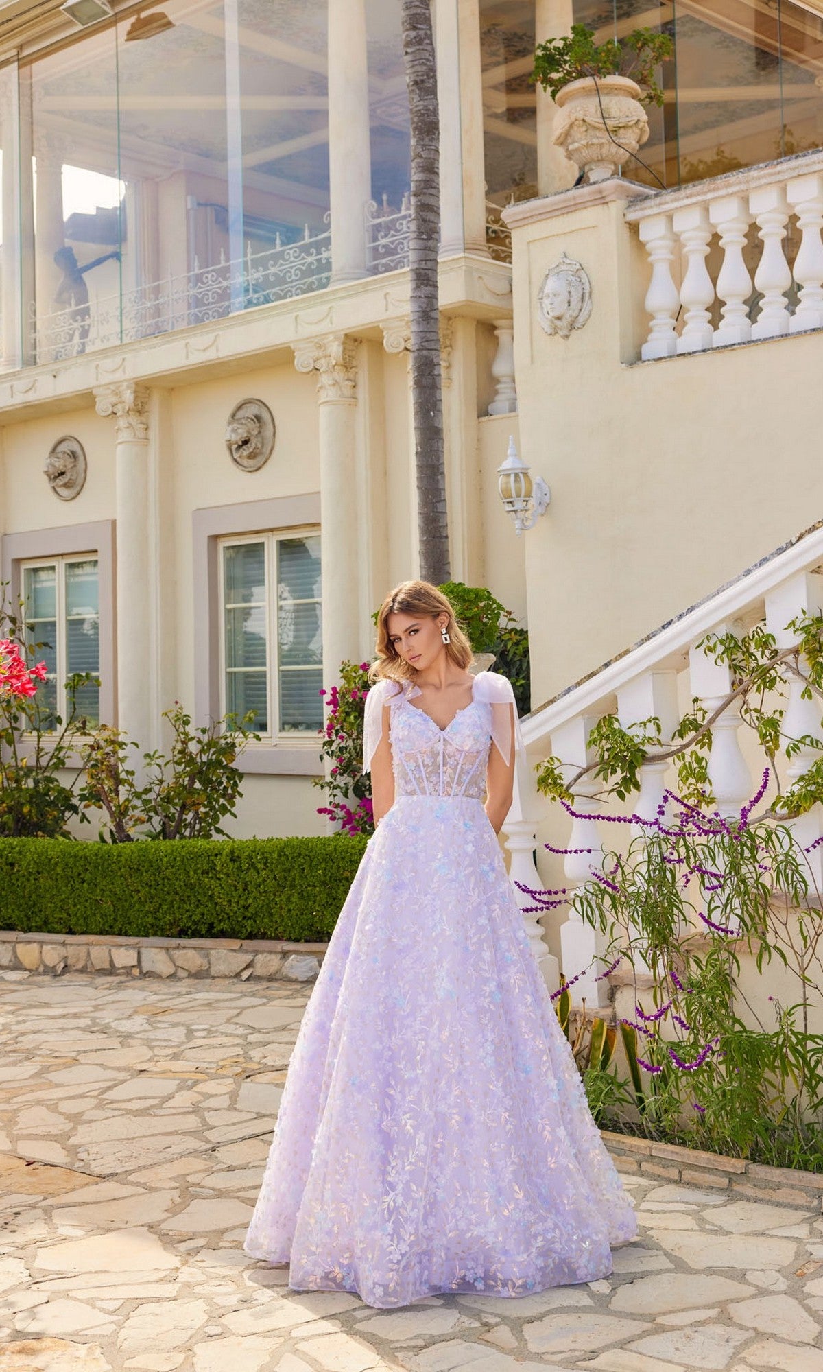 A woman in the Juliet JT2439K Lilac Purple Long Lace Prom Dress stands outdoors before a cream-colored, ornate building with columns and a balcony.