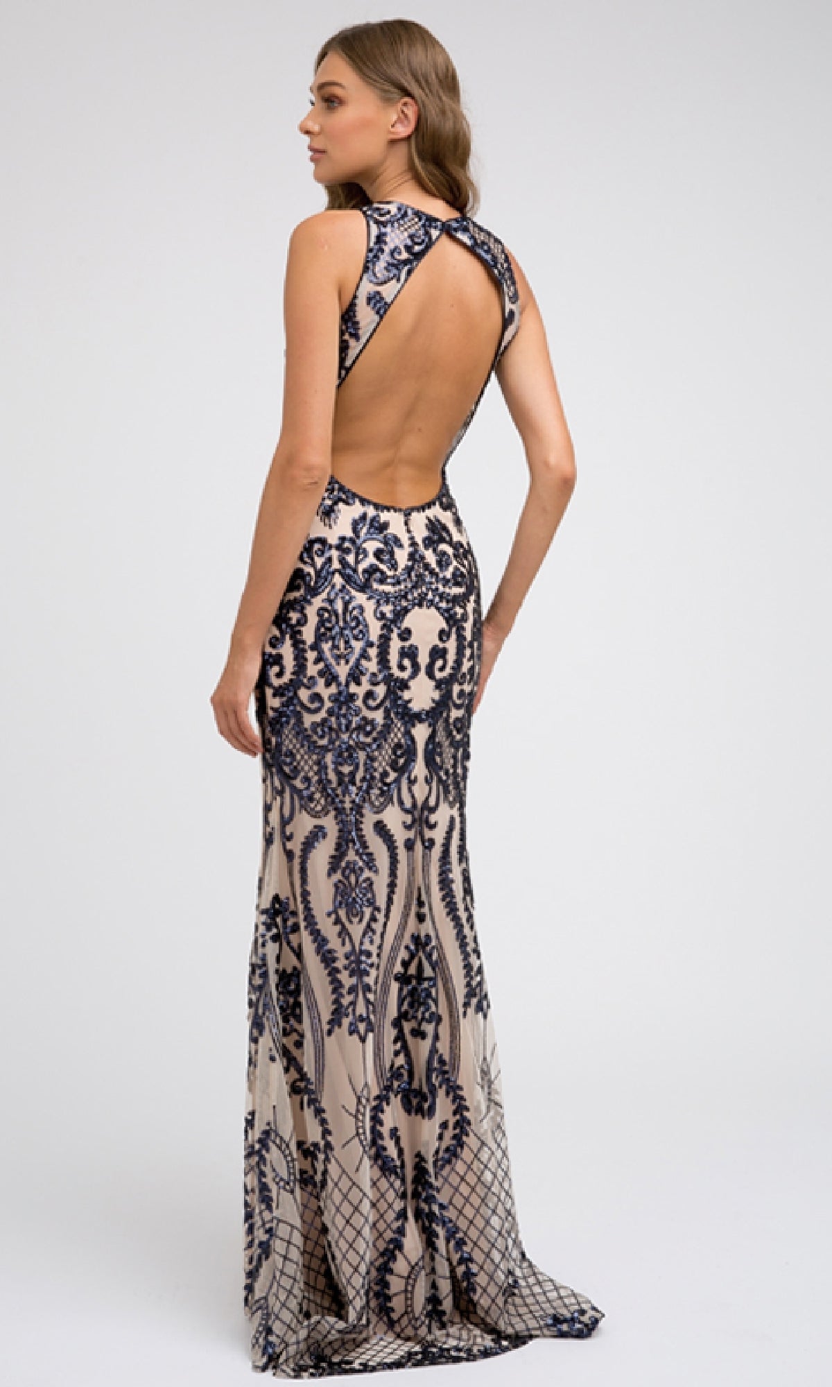 A woman models the Juliet 243 long prom dress, featuring a sleeveless, open-back design with intricate navy blue embroidery on light beige fabric, set against a plain background.