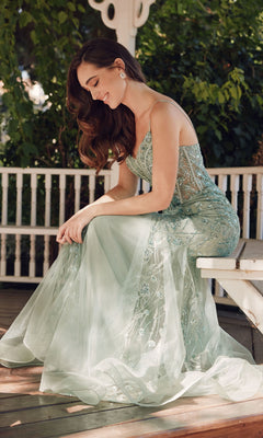 A woman in the Juliet JT2441S Long Sequin Mermaid Prom Dress sits on a white bench outdoors, smiling and looking down, surrounded by greenery and a white wooden railing.