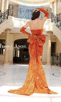 A woman wears the Gloved Short Sequin Hoco Dress: Jessica Angel 2452—an orange strapless mini dress with a large bow and long train—in an elegant room with columns and a balcony. Text reads "Jessica Angel Made in USA. Style: 2452.