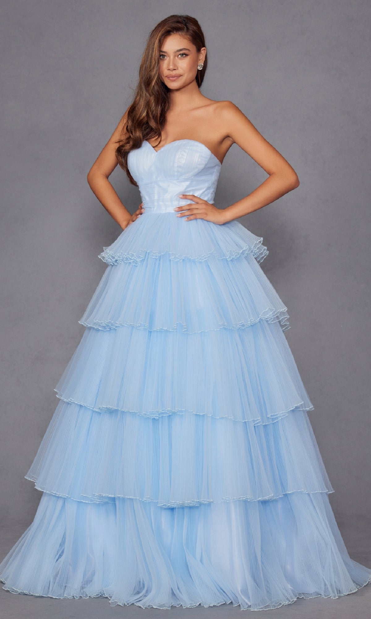 A woman stands with hands on hips wearing the Strapless Blue Prom Ball Gown: Juliet JT2452K, a long, tiered, ruffled and elegant dress, against a plain gray background.