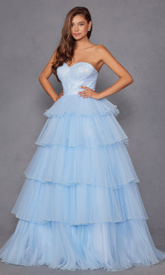 A woman stands with hands on hips wearing the Strapless Blue Prom Ball Gown: Juliet JT2452K, a long, tiered, ruffled and elegant dress, against a plain gray background.