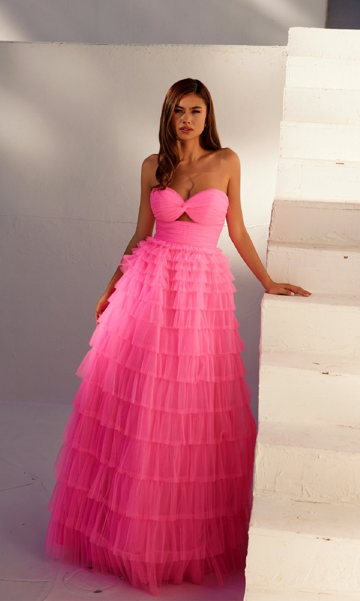 A woman in the Long Prom Dress JT2456H by Juliet, featuring bright pink ruffle tulle and an empire waist, stands beside a white painted staircase.