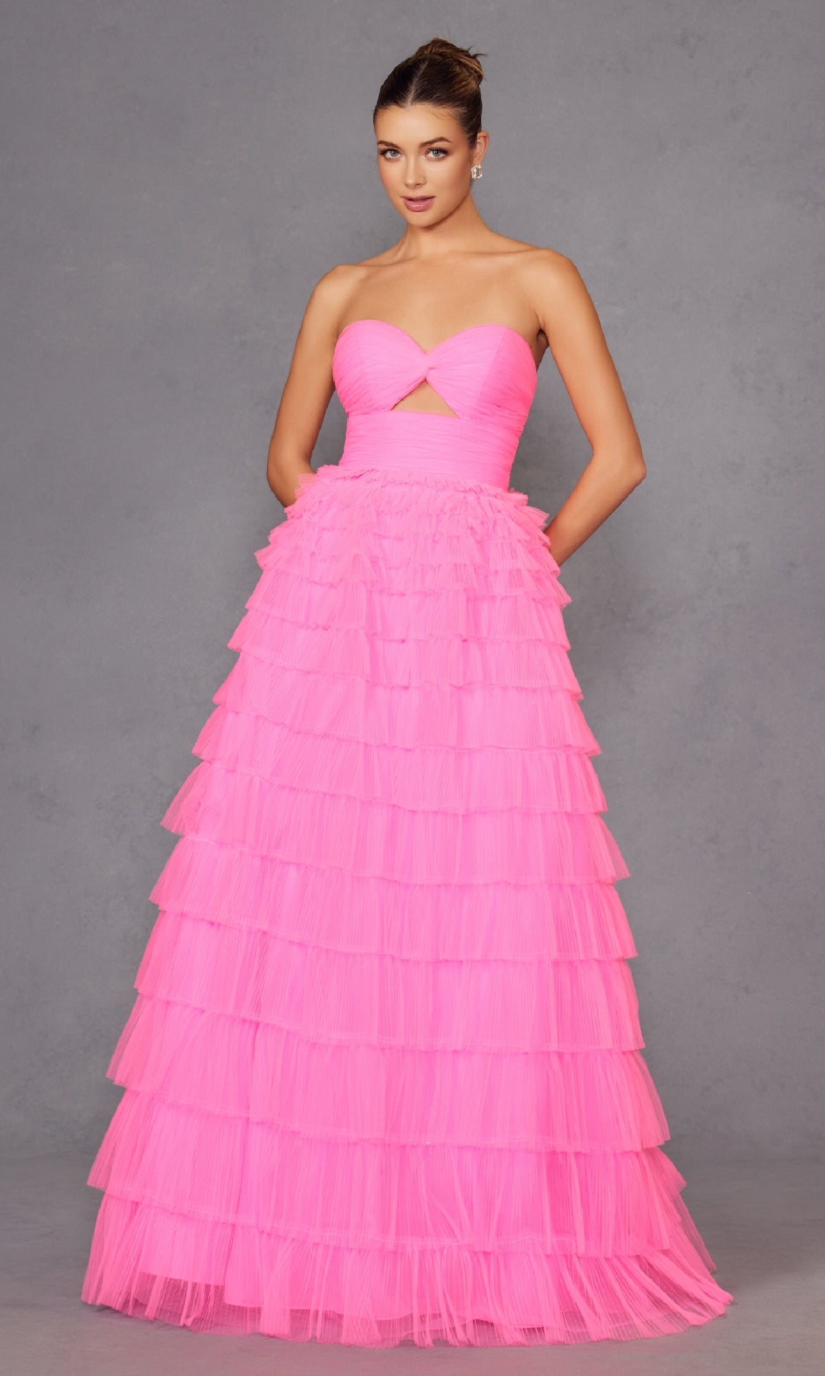 A woman stands against a gray background wearing the Long Prom Dress JT2456H by Juliet, a strapless ball gown with an empire waist, floor-length tiered bright pink ruffle tulle skirt, and a small bust cutout.