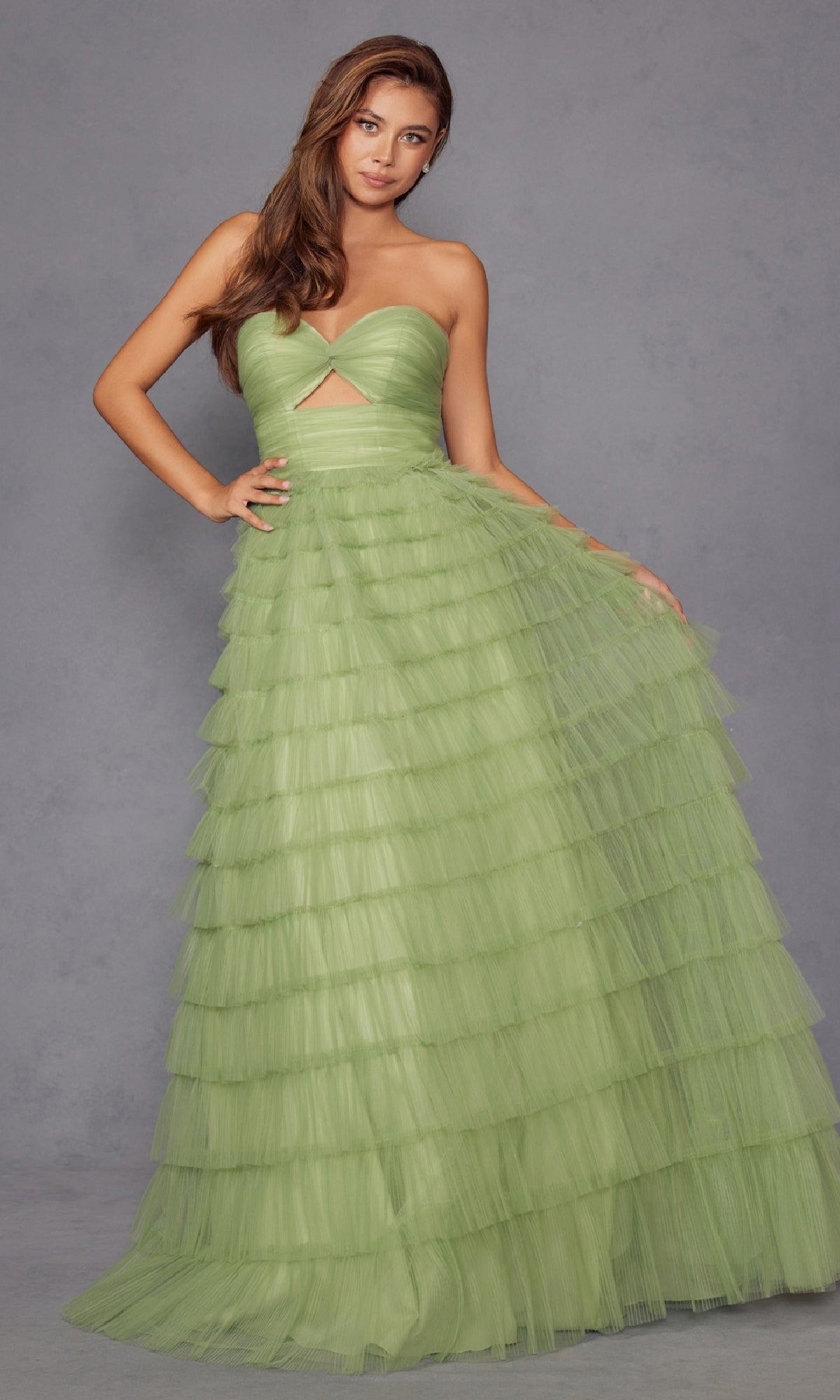 A woman stands against a plain gray backdrop wearing the Long Prom Dress JT2456H by Juliet, a strapless light green ruffle tulle ball gown with an empire waist and a triangular midriff cutout.
