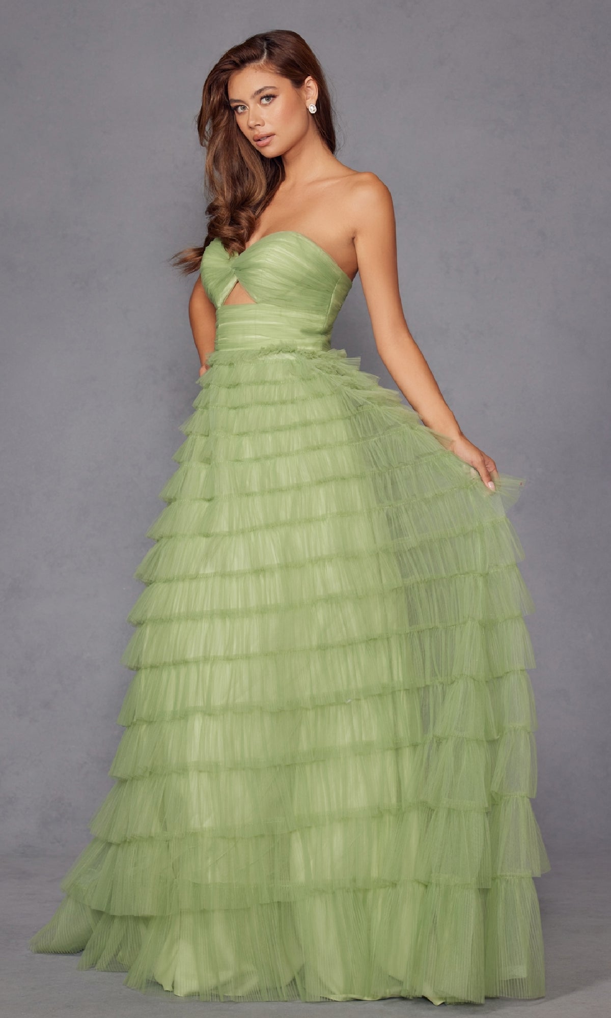 A woman in the Long Prom Dress JT2456H by Juliet, a light green strapless ball gown with ruffled tulle, tiered skirt, and waist cutout, stands against a plain gray background.