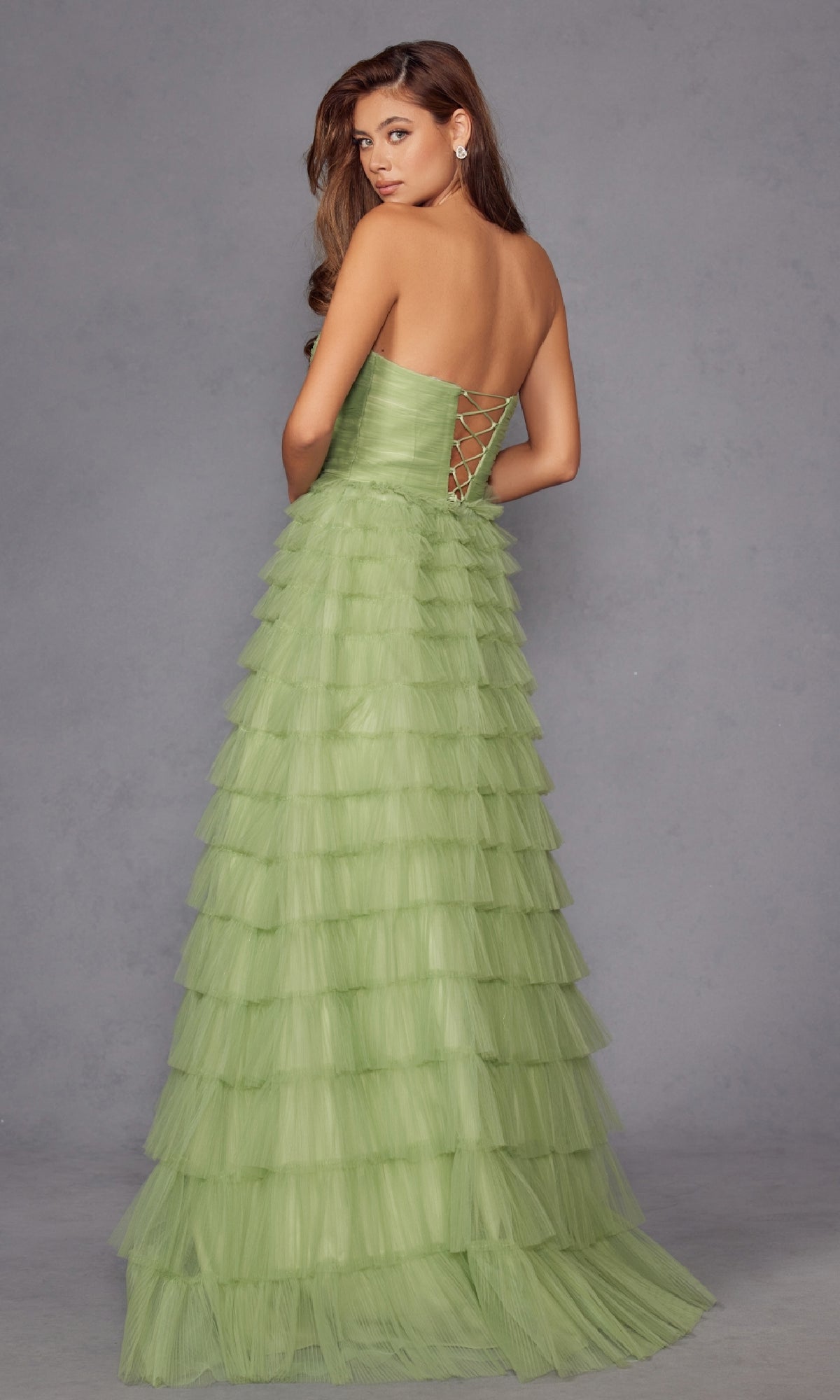 A woman stands against a plain gray background, facing away and looking over her shoulder, wearing the Long Prom Dress JT2456H by Juliet—a strapless ball gown with light green ruffle tulle and an empire waist.
