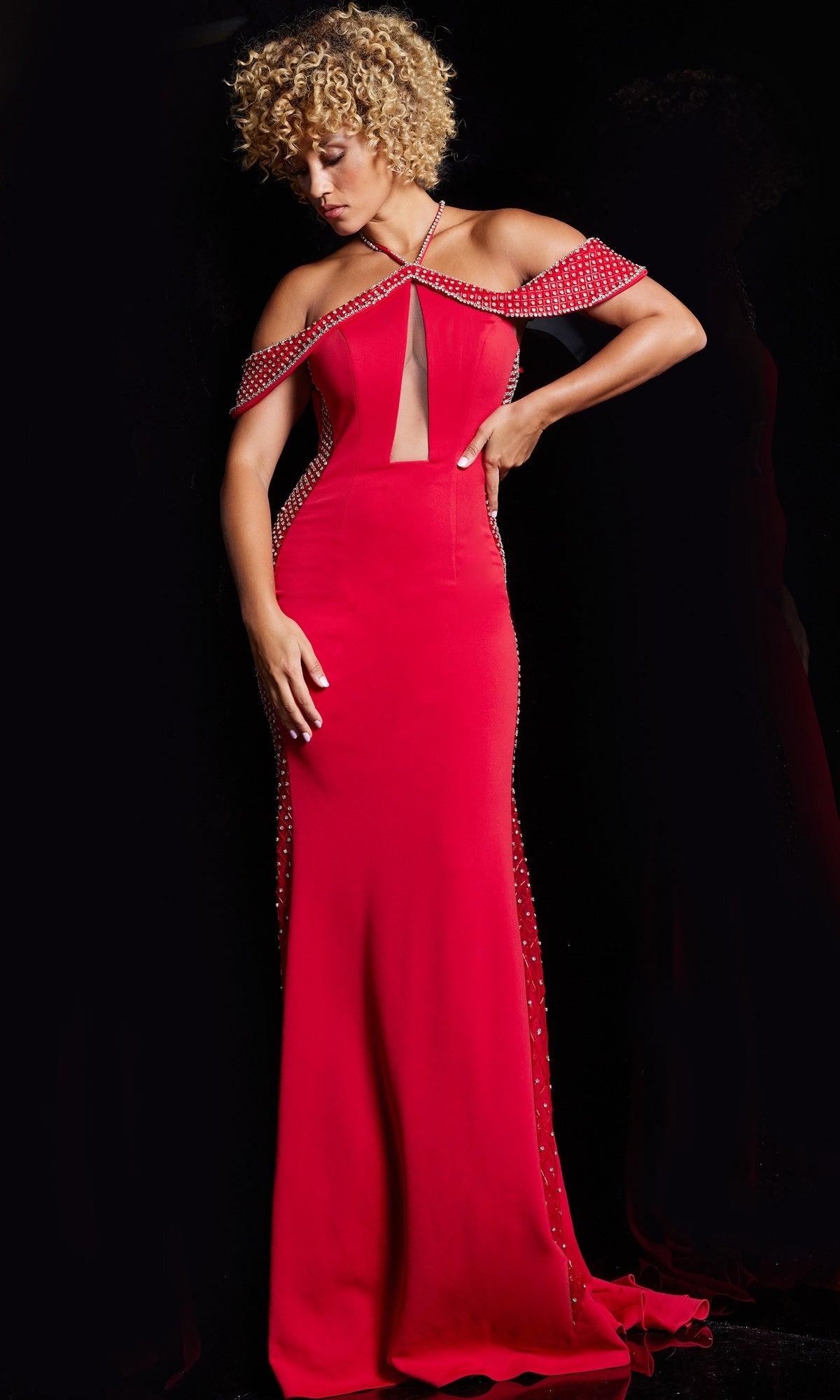 A woman with curly blonde hair wears the Jovani Sheer-Illusion-Sides Long Prom Dress 24611 in bright red, featuring beaded embellishments, cold shoulder cutouts, and a front slit, standing against a black background.