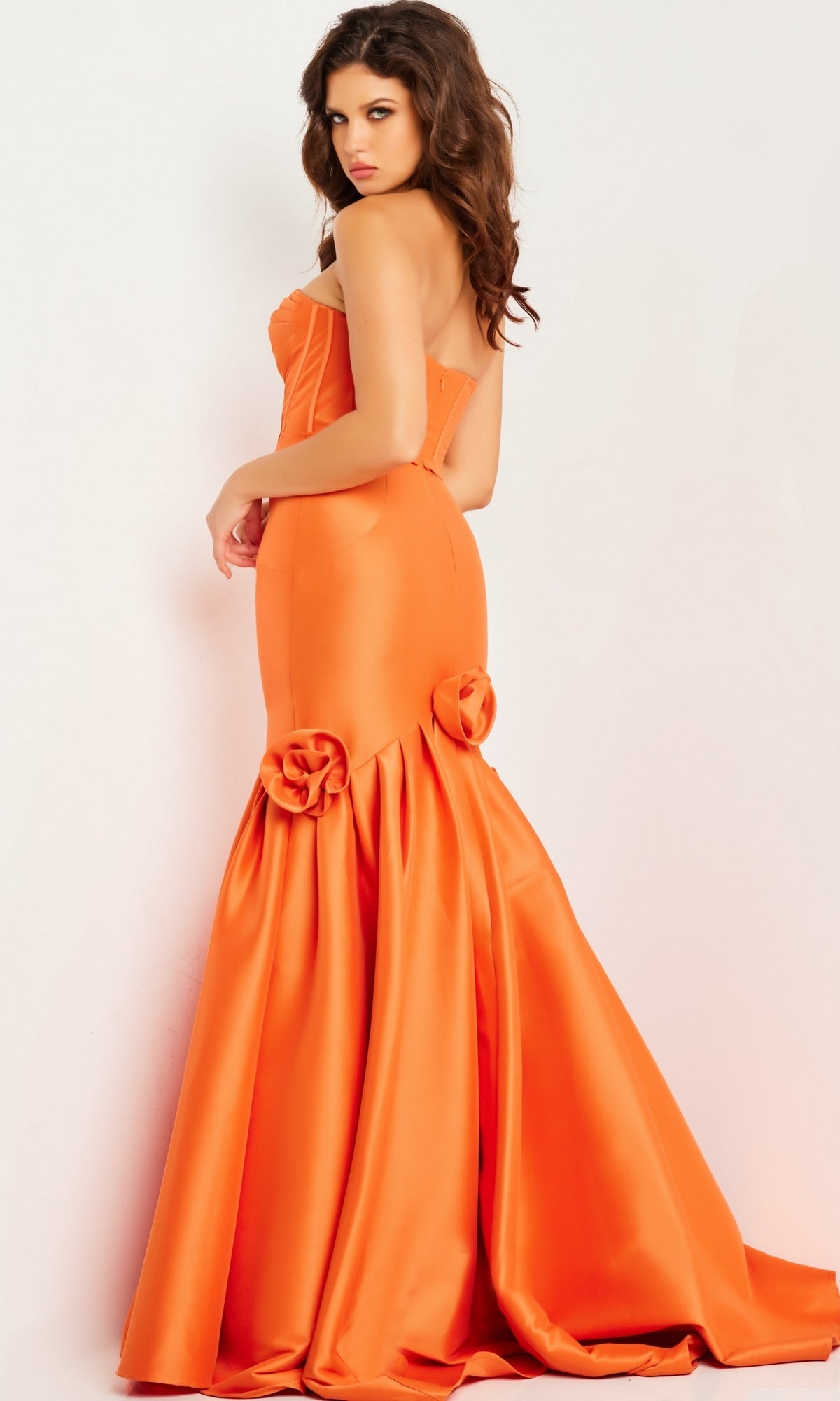 Woman wearing the Jovani Strapless Long Mermaid Prom Dress 24613 in orange satin, featuring a fitted silhouette, flared skirt, and rose fabric details at the knees, posing against a plain white background.