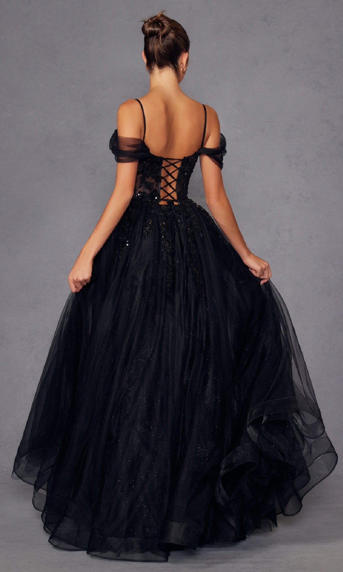 A woman stands with her back to the camera, showcasing the Juliet JT2466A Sheer-Waist Long Prom Ball Gown—an elegant black off-the-shoulder A-line dress with a lace-up corset back and full tulle skirt, perfect for any formal occasion.