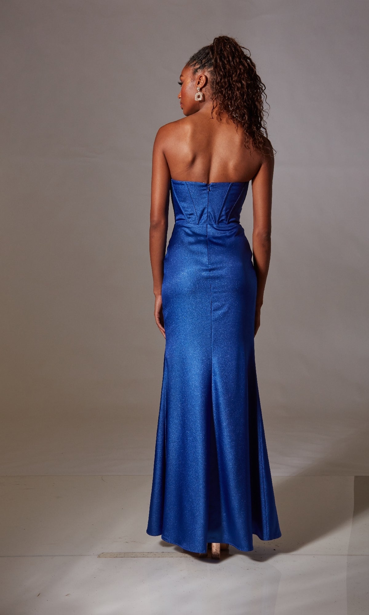 A woman, facing away, wears the Strapless Long Prom Dress: Aerie Couture 246—a blue, fitted, floor-length gown—paired with hoop earrings against a plain background.
