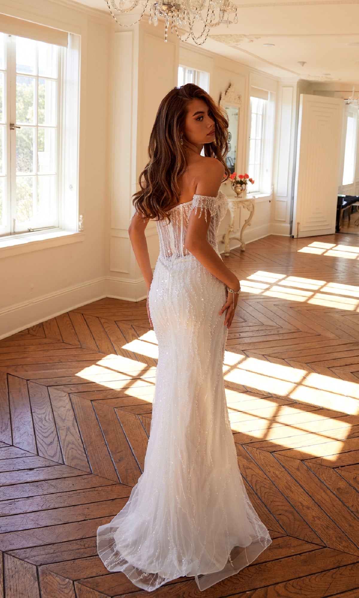 In a sunlit room with wooden floors, a woman stands facing away, showcasing the elegant silhouette of the Long Prom Dress JT2471LW by Juliet—a fitted, white sequin off-shoulder gown.