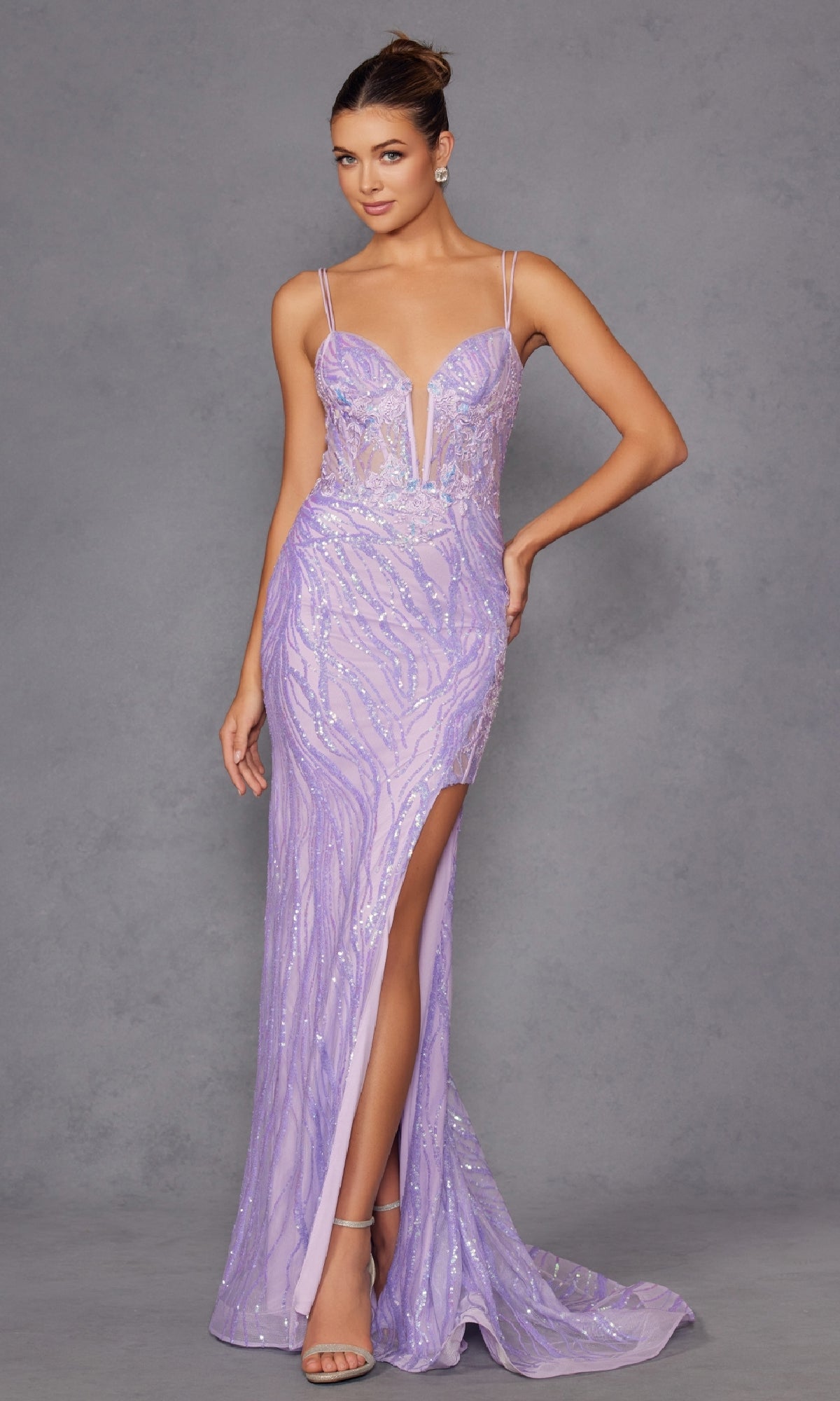 A woman in the Juliet JT2473R Long Lilac Sequin Prom Dress with thin straps, a deep neckline, and a thigh-high slit stands against a gray background.