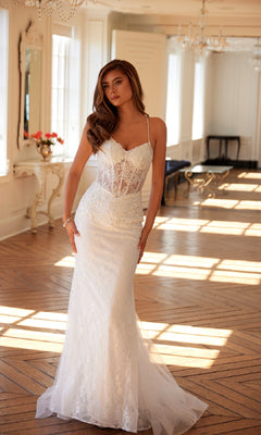 A woman in the Long White Bridal Dress: Juliet JT2481UW, featuring off-white lace, thin straps, and a sheer corset bodice, stands in a sunlit elegant room with large windows and wooden floors.