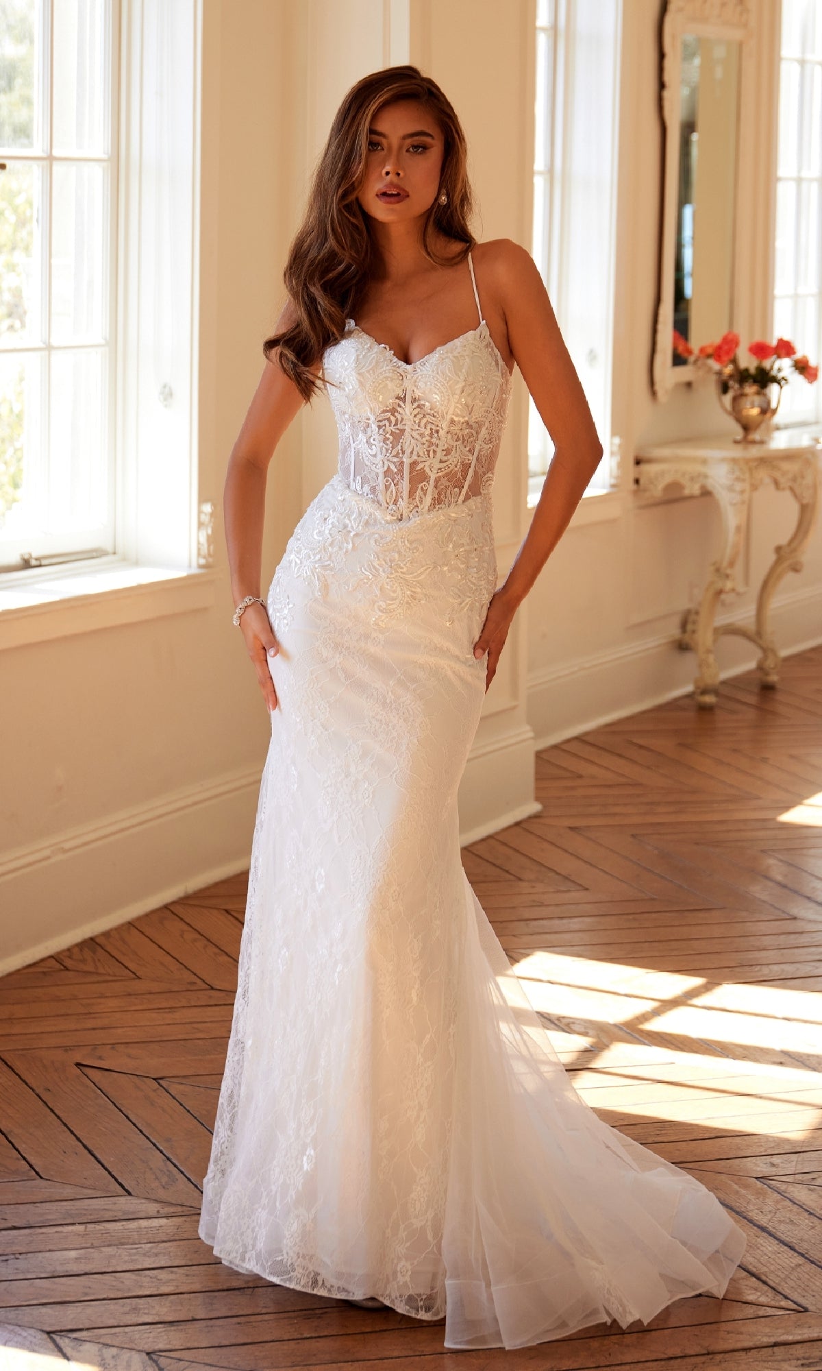 A woman stands indoors wearing the Long White Bridal Dress: Juliet JT2481UW, featuring thin straps, an off-white lace design with a sheer corset bodice, and delicate floral lace embroidery.