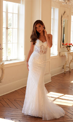 A woman stands indoors in the Long White Bridal Dress: Juliet JT2481UW, an off-white gown with a fitted bodice, flowing train, and floral lace embroidery, surrounded by large windows and elegant decor.