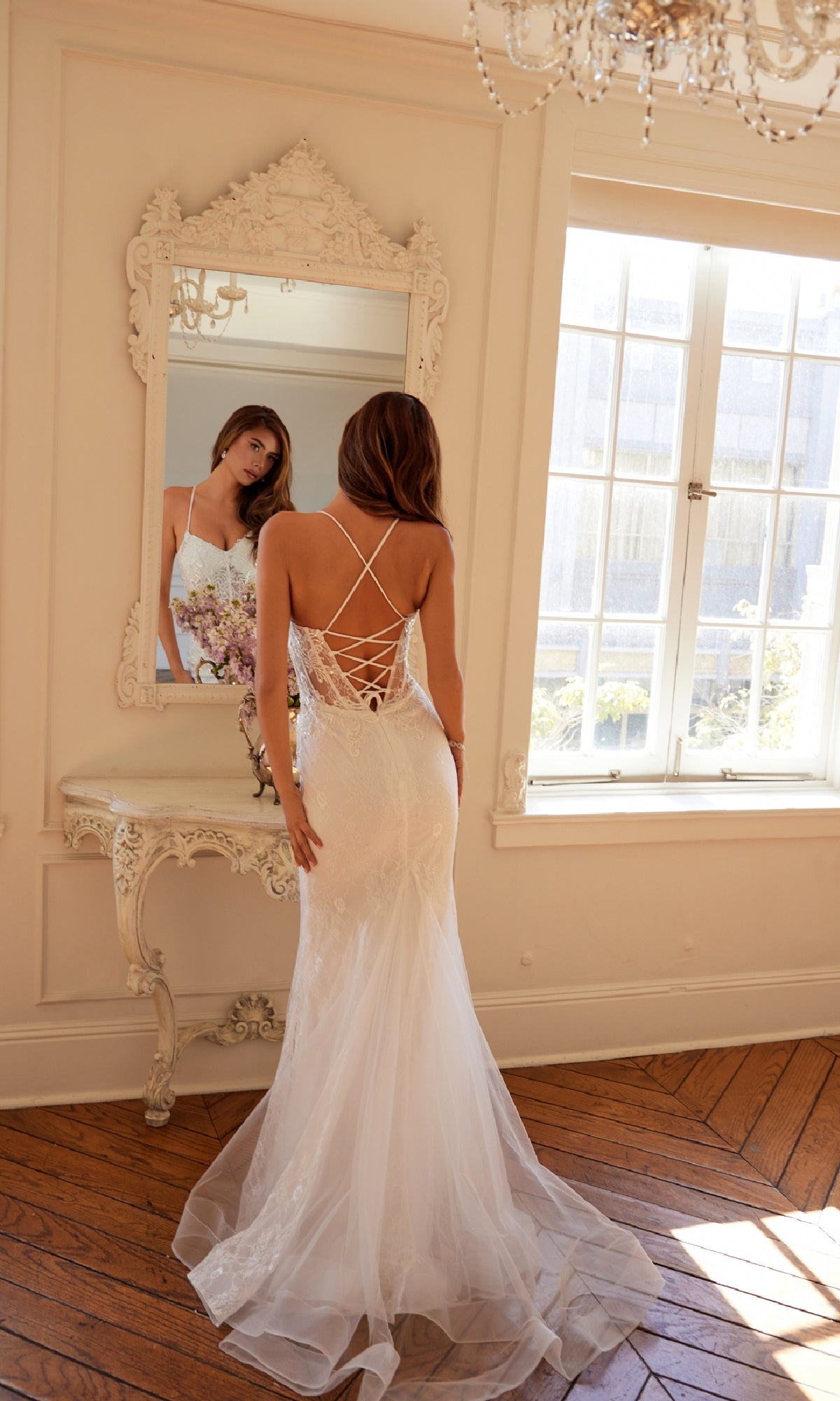 A woman wearing the Long White Bridal Dress: Juliet JT2481UW with an off-white lace finish and sheer corset bodice stands before a mirror in a bright, ornately decorated room with wooden floors.