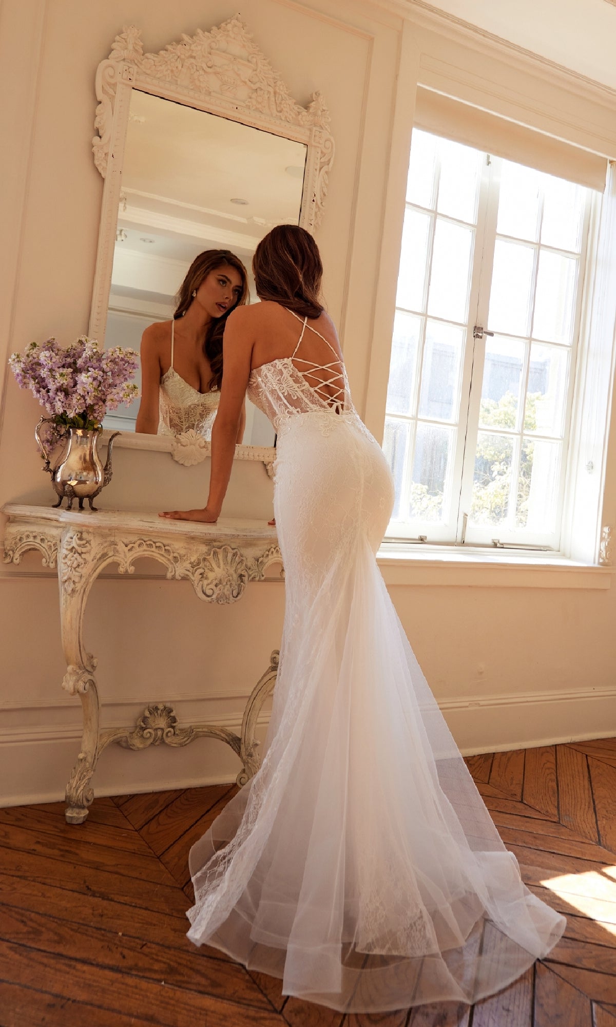 A woman wearing the Long White Bridal Dress: Juliet JT2481UW leans over an ornate vanity, gazing at her reflection in a sunlit room.