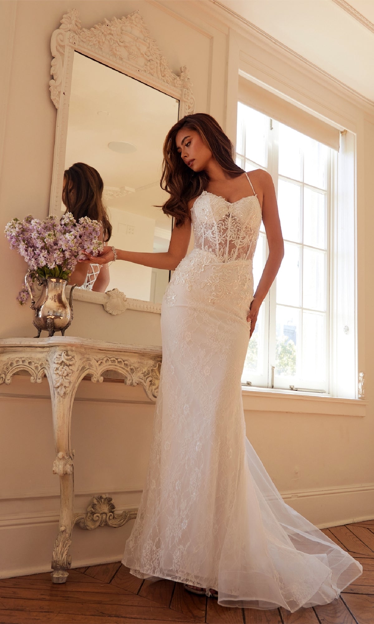 A woman in the Juliet JT2481UW long white bridal dress with a sheer corset bodice stands by an ornate mirror, holding a vase of lilac flowers in a sunlit room with wooden floors.
