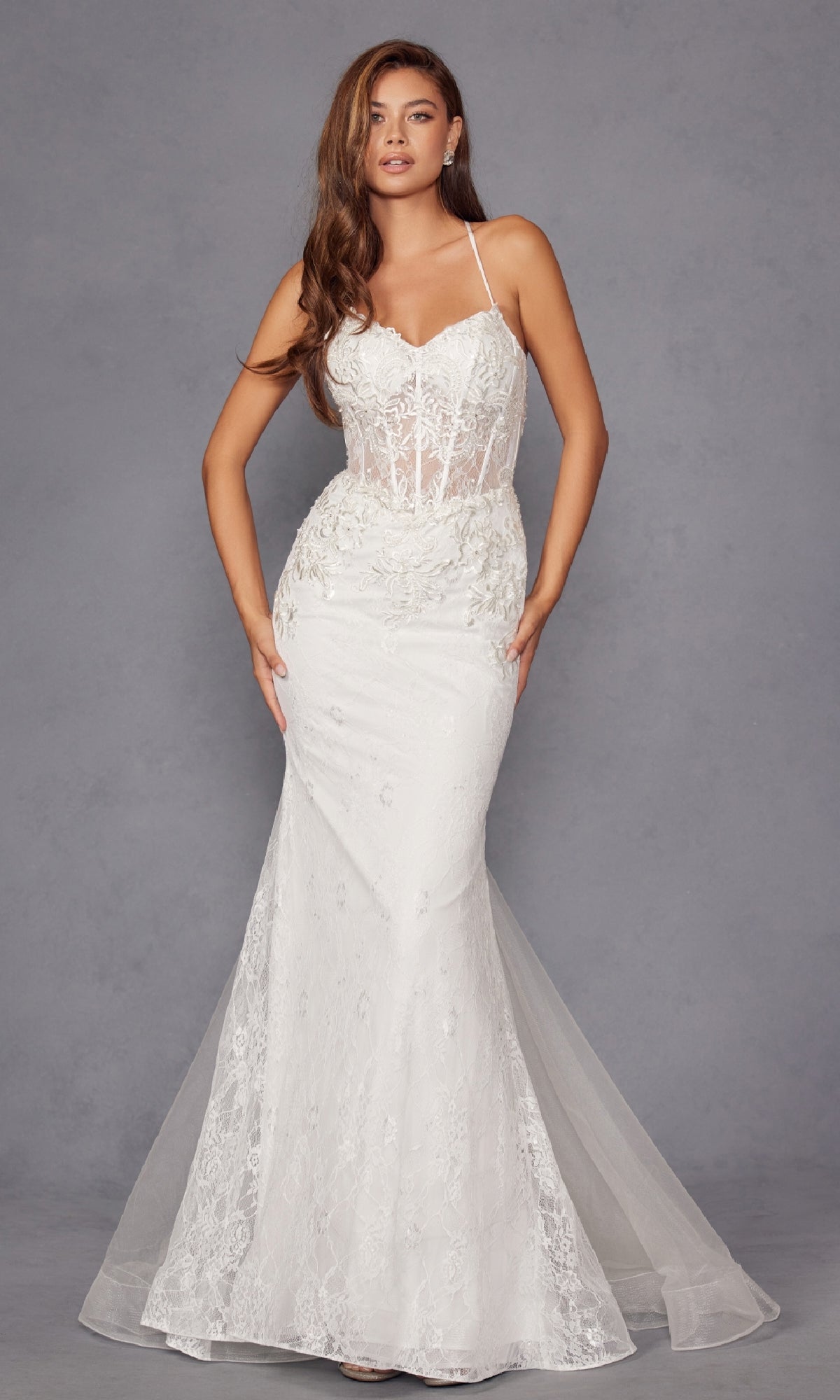 A woman wears the Long White Bridal Dress: Juliet JT2481UW, a fitted off-white gown with thin straps, sheer corset bodice, and floral lace embroidery, standing against a plain gray background.